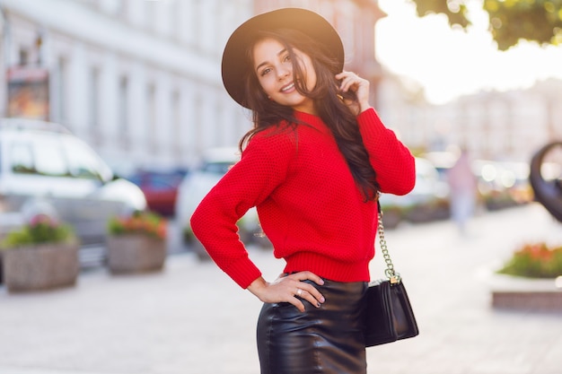 Foto gratuita imagen de estilo callejero de moda al aire libre de mujer morena seductora en otoño casual traje caminando en ciudad soleada. jersey de punto rojo, sombrero de moda negro, falda de cuero.