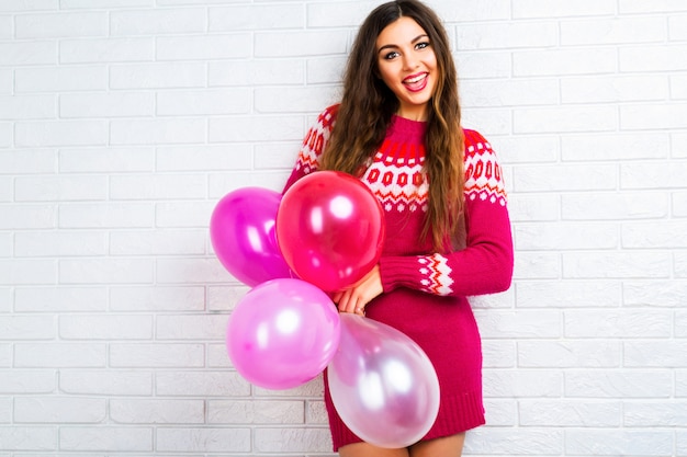 Imagen de estilo de vida interior de una divertida chica morena bonita con maquillaje brillante y pelos largos, vistiendo un suéter moderno y sosteniendo globos de fiesta rosa.