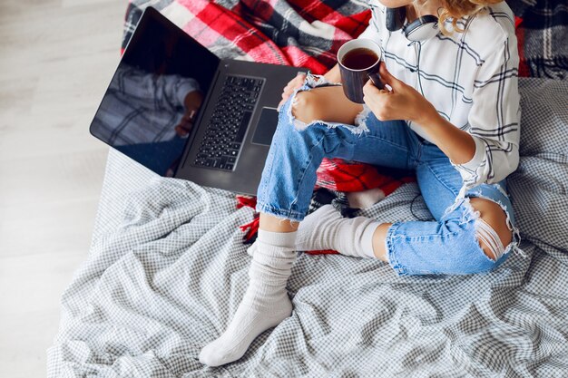 Imagen de estilo de vida, mujer tomando café y usando la computadora, vistiendo calcetines calientes y jeans de moda. Sentado en la cama. Temprano en la mañana. Vista superior.