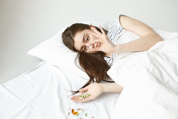 Imagen de una estudiante de cabello oscuro que pasa el día en la cama, tratando de recuperarse de la gripe, sosteniendo un montón de pastillas de colores en las manos y se derrama sobre una sábana blanca, eligiendo cuál tener para curarse