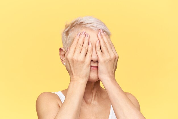 Imagen de estudio aislado de abuela irreconocible con cabello rubio corto jugando al escondite con sus nietos, cubriéndose los ojos con ambas manos.