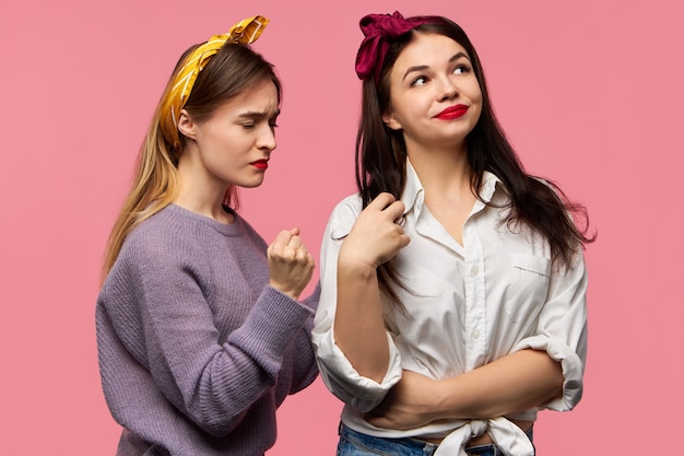 Imagen de estudio de indignada joven mujer caucásica enojada perdiendo los estribos estando enojada con su amiga mala que no le importa en absoluto. Dos mujeres que expresan diferentes emociones, posando aisladas