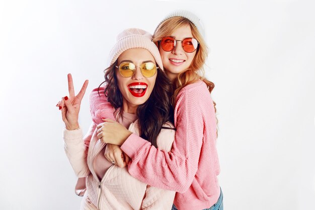 Imagen de estudio interior de dos niñas, amigos felices con ropa rosa con estilo y sombrero gracioso deletrear juntos. Fondo blanco. Sombrero y gafas de moda. Mostrando paz.