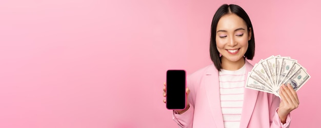 Imagen de una exitosa mujer corporativa coreana que muestra dinero en dólares y la interfaz de pantalla de la aplicación de teléfono inteligente del concepto de aplicación de teléfono móvil de fondo rosa de inversión y finanzas