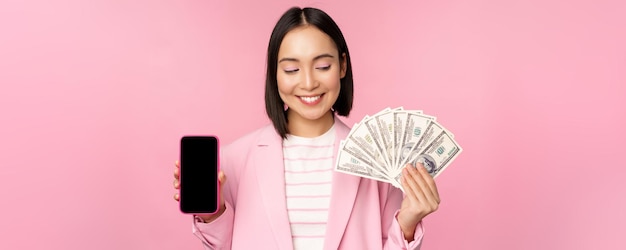 Imagen de una exitosa mujer corporativa coreana que muestra dinero en dólares y la interfaz de pantalla de la aplicación de teléfono inteligente del concepto de aplicación de teléfono móvil de fondo rosa de inversión y finanzas