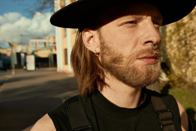 Imagen exterior de apuesto joven sin afeitar con aretes, piercing en la nariz y sombrero negro caminando por las calles de la ciudad, disfrutando de un día soleado