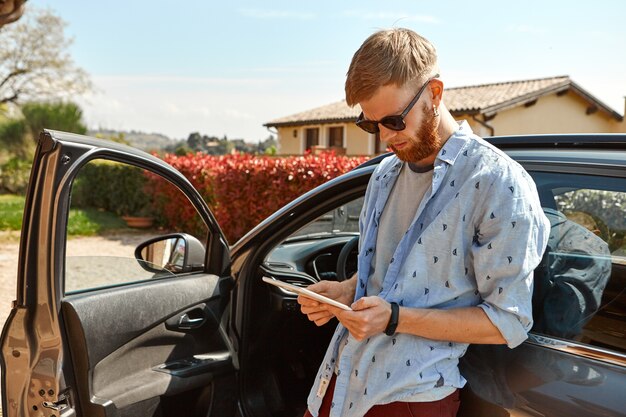 Imagen exterior de chico guapo hipster de moda con barba borrosa de pie en su coche