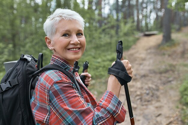 Imagen exterior de una hermosa mujer madura enérgica con mochila con bastones, disfrutando de nordic walking en el bosque, mirando a la cámara con una sonrisa feliz