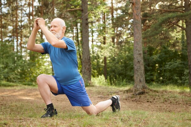 Imagen exterior de un hombre mayor activo en zapatillas para correr dando un paso hacia adelante haciendo estocadas, manteniendo las manos juntas frente a su cara. Atractivo pensionista masculino sano estirando los músculos de las piernas en el bosque