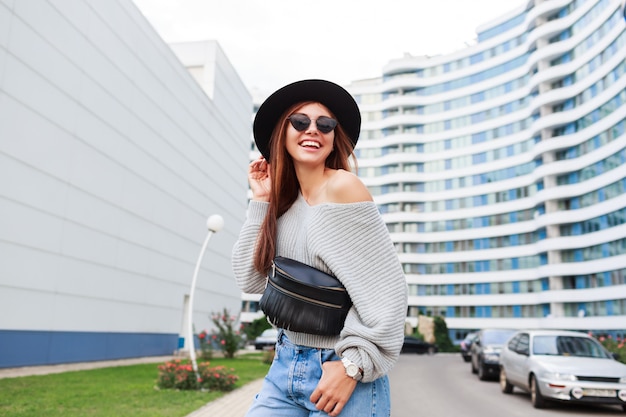 Foto gratuita imagen exterior de niña alegre con sombrero de lana negra y suéter de otoño gris saltando y disfrutando de caminar en la ciudad urbana moderna.