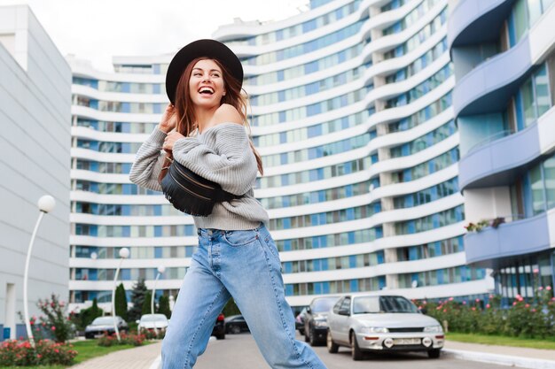 Imagen exterior de niña alegre con sombrero de lana negra y suéter de otoño gris saltando y disfrutando de caminar en la ciudad urbana moderna.