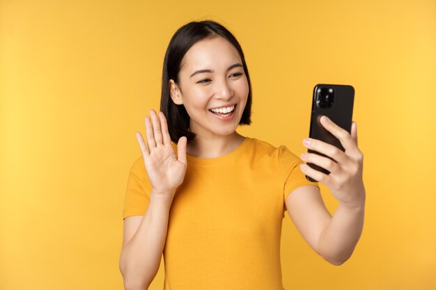 Imagen de feliz hermosa chica asiática chateando en video hablando en una aplicación de teléfono inteligente de pie contra un fondo amarillo