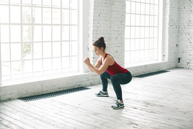 Imagen de fuerte chica deportiva vistiendo elegante camiseta sin mangas, zapatillas y leggings haciendo sentadillas sobre un piso de madera en el centro de gimnasio contra grandes ventanales