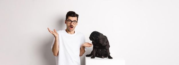 Foto gratuita imagen de un gracioso pug negro mirando a su confundido dueño encogiéndose de hombros desconcertado de pie sobre el bac blanco