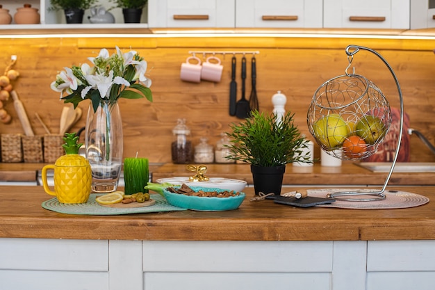 Imagen de la gran cocina luminosa con alacenas blancas y marrones con tetera de piña amarilla, molinillo de pimienta blanca y colgante de metal con frutas y galletas