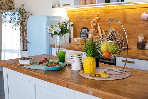 Imagen de la gran cocina luminosa con alacenas blancas y marrones con tetera de piña amarilla, molinillo de pimienta blanca y colgantes de metal con frutas