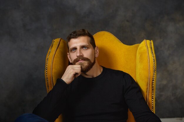 Imagen de guapo joven caucásico barbudo con elegante suéter negro relajándose en un lujoso sillón amarillo, manteniendo la mano en la barbilla, reflexionando, con expresión pensativa y pensativa