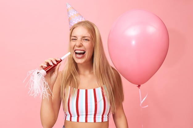 Imagen de hermosa adolescente linda con cabello rubio suelto y tirantes posando en rosa con soplador de fiesta y globo de helio, riendo con la boca abierta