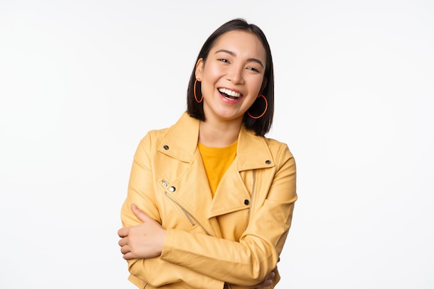 Imagen de una hermosa chica asiática moderna riéndose sonriendo y mirando feliz a la cámara de pie en una chaqueta amarilla contra el concepto de personas de fondo blanco