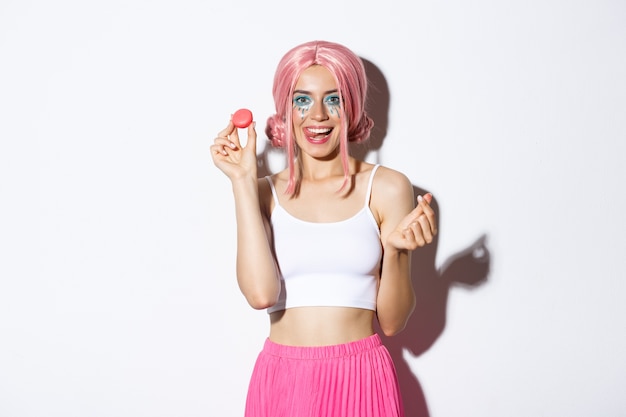 Imagen de hermosa chica con maquillaje brillante comiendo sabrosos macarrones, con peluca rosa, mostrando la lengua y sonriendo, de pie.