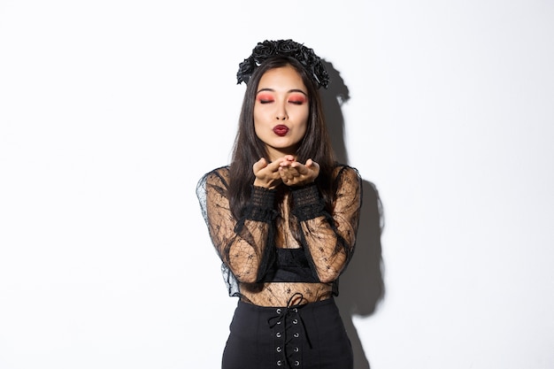 Imagen de hermosa y elegante mujer asiática en traje de bruja, cerrar los ojos y enviar aire beso t cámara, con vestido gótico con corona negra, de pie sobre fondo blanco.