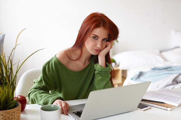 Imagen de una hermosa estudiante europea vestida de manera informal con cabello rojo con expresión aburrida mientras realiza un proyecto de investigación en casa, sentada en el lugar de trabajo con computadora portátil, teléfono y libros de texto en el escritorio