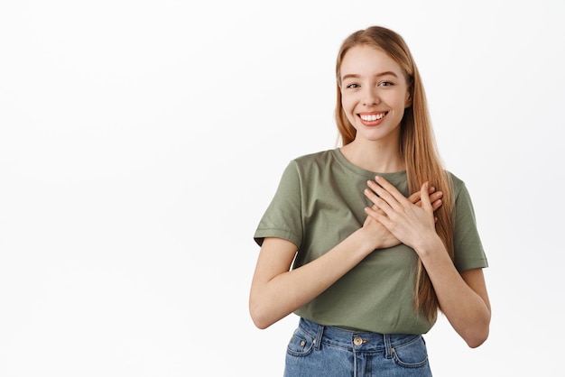 Imagen de una hermosa joven cogida de la mano en el corazón sonriendo y luciendo complacida halagada recibir una sincera sorpresa siendo tocada y encantada de pie sobre fondo blanco