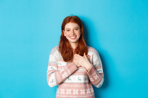 Imagen de la hermosa modelo femenina pelirroja tomados de la mano en el corazón y sonriendo, diciendo gracias, siendo agradecido, de pie sobre fondo azul