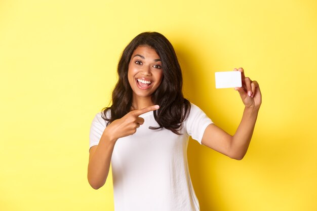 Imagen de hermosa mujer afroamericana en camiseta blanca apuntando a la tarjeta de crédito y sonriendo