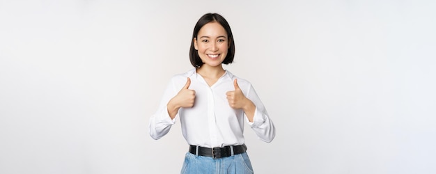 Imagen de una hermosa mujer asiática adulta que muestra los pulgares hacia arriba usando ropa formal de la universidad de la oficina recomendando compañía de pie sobre fondo blanco