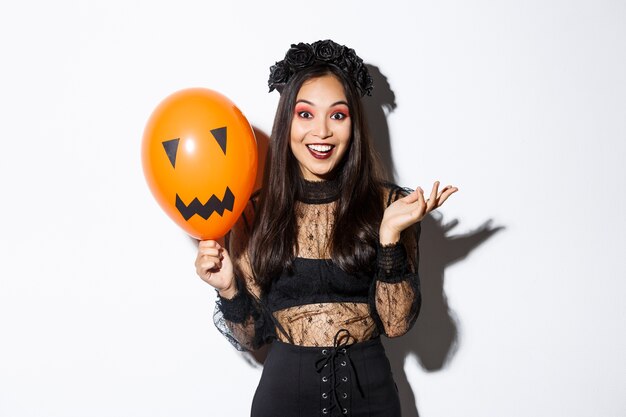 Imagen de hermosa mujer asiática celebrando halloween, vestida con traje de bruja y maquillaje gótico, hablando con globo naranja.