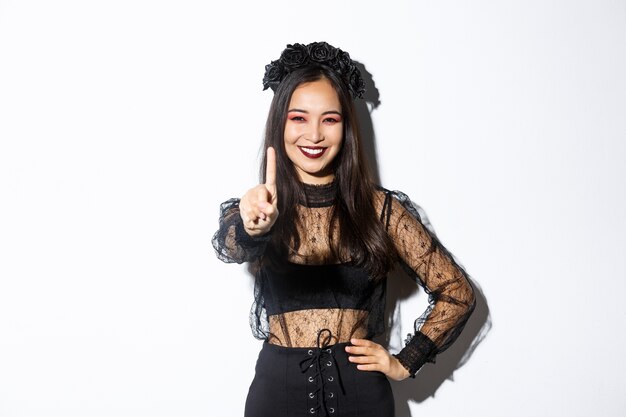 Imagen de una hermosa mujer asiática sonriente en traje de halloween que muestra un gesto de parada, extiende un dedo y parece feliz, prohíbe o rechaza algo, fondo blanco de pie.