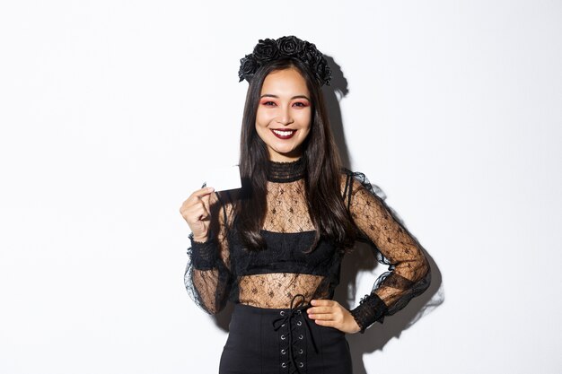 Imagen de hermosa mujer asiática en traje de halloween, mostrando tarjeta de crédito y sonriendo, de pie con vestido de encaje gótico sobre fondo blanco.