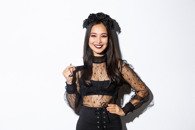 Imagen de hermosa mujer asiática en traje de halloween, mostrando tarjeta de crédito y sonriendo, de pie con vestido de encaje gótico sobre fondo blanco.