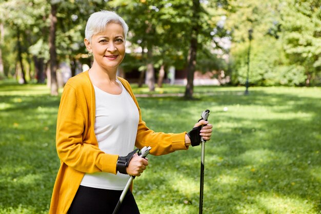 Imagen de hermosa mujer madura anciana alegre en cardigan amarillo sosteniendo palos para caminata nórdica, disfrutando de un estilo de vida activo y saludable, sintiéndose lleno de energía, con una sonrisa feliz