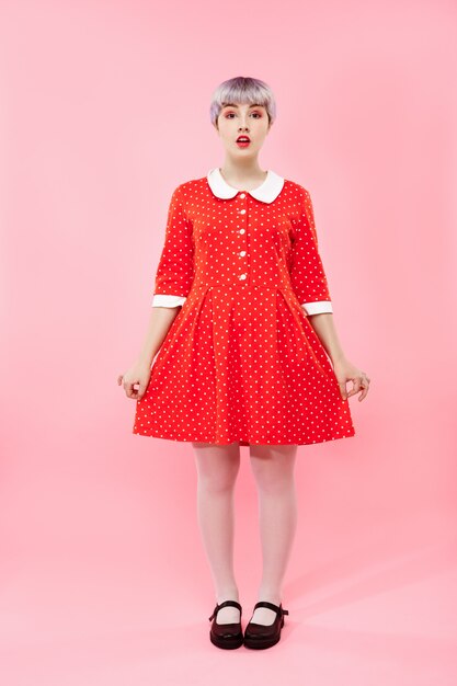 Imagen de una hermosa niña dollish con cabello corto violeta claro con un vestido rojo sobre una pared rosa