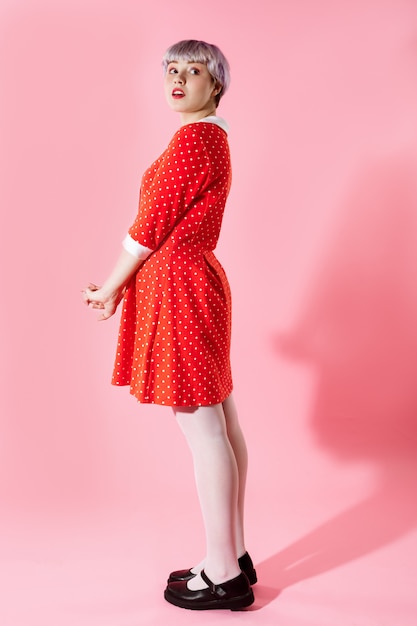 Imagen de una hermosa niña dollish con cabello corto violeta claro con un vestido rojo sobre una pared rosa