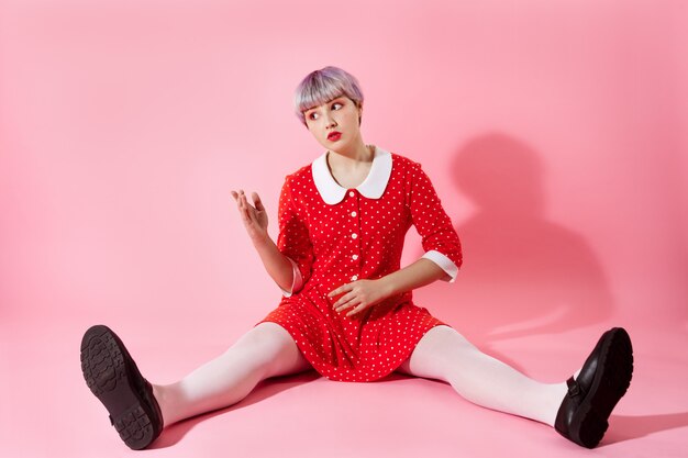 Imagen de una hermosa niña dollish con cabello corto violeta claro con un vestido rojo sobre una pared rosa