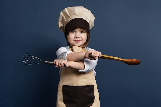 Imagen de una hermosa niña con gorro de chef y delantal sosteniendo una batidora de mano o un batidor de alambre en una mano y una cuchara de madera en la otra, yendo a batir huevos o hacer salsa de tomate. Concepto de comida y cocina