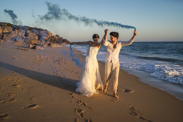 Imagen de una hermosa pareja posando con una bomba de humo azul en la playa