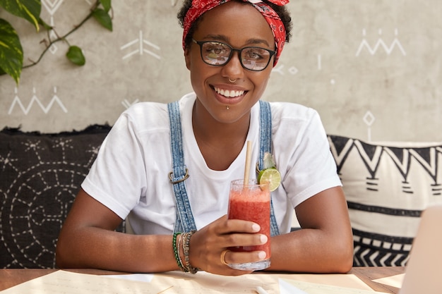 Imagen de hipster alegre lleva gafas transparentes, vestida con ropa casual, bebidas cócteles frescos