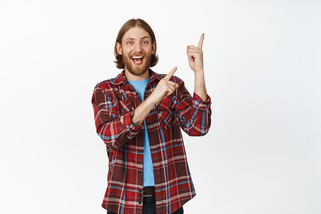 Imagen de un hipster feliz, un hombre rubio barbudo que muestra un banner, haga clic en el enlace, señalando con el dedo en la esquina superior derecha y sonriendo emocionado de pie sobre un fondo blanco