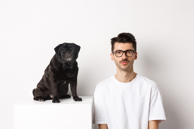 Imagen de hipster guapo con gafas sentado junto al perro pug lindo negro, ambos mirando a cámara sobre fondo blanco.