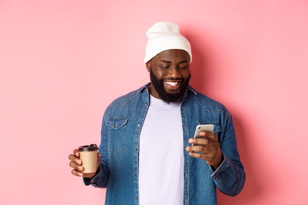 Imagen de hipster hombre negro elegante bebiendo café para llevar, leyendo un mensaje en el teléfono y sonriendo, de pie sobre fondo rosa.