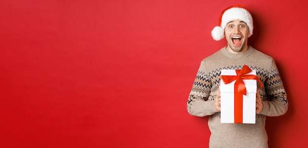 Imagen de un hombre apuesto y emocionado que recibe un regalo de Navidad, usa un sombrero de Papá Noel y un suéter de invierno, grita de alegría, sostiene un regalo y se para sobre un fondo rojo.