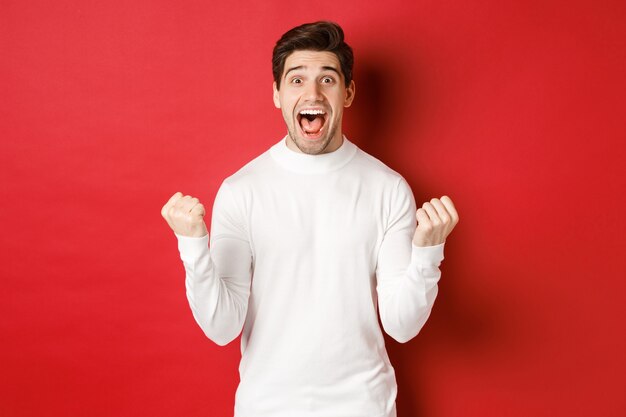 Imagen de hombre apuesto feliz en suéter blanco ganando algo haciendo bomba de puño y sonriendo asombro ...