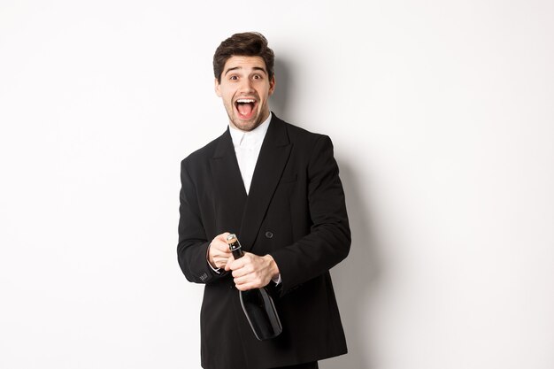 Imagen de hombre atractivo en traje negro con una fiesta, celebrando el año nuevo y abriendo una botella de champán, de pie feliz contra el fondo blanco.