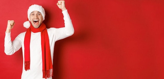 Imagen de hombre caucásico alegre con gorro de Papá Noel y bufanda, gritando de alegría y levantando las manos, celebrando la victoria o la victoria, triunfando sobre fondo rojo