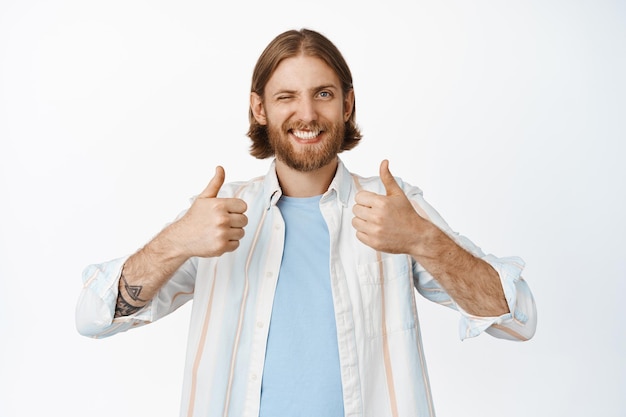 Foto gratuita la imagen de un hombre caucásico alegre tiene una opinión positiva, me gusta y está de acuerdo, guiñando un ojo y sonriendo feliz, mostrando el pulgar hacia arriba en aprobación, de pie sobre fondo blanco.