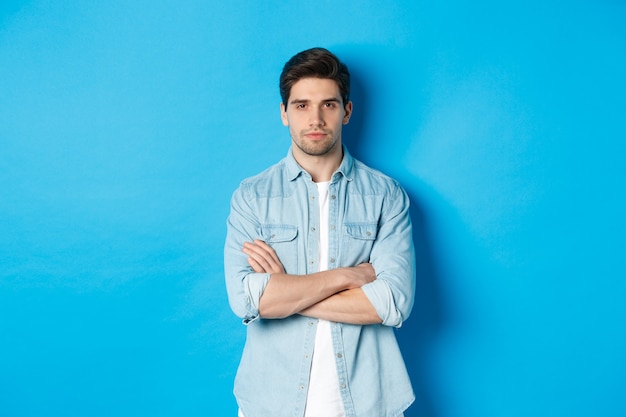 Imagen de hombre caucásico guapo en traje casual, mirando serio y seguro, de pie contra el fondo azul.